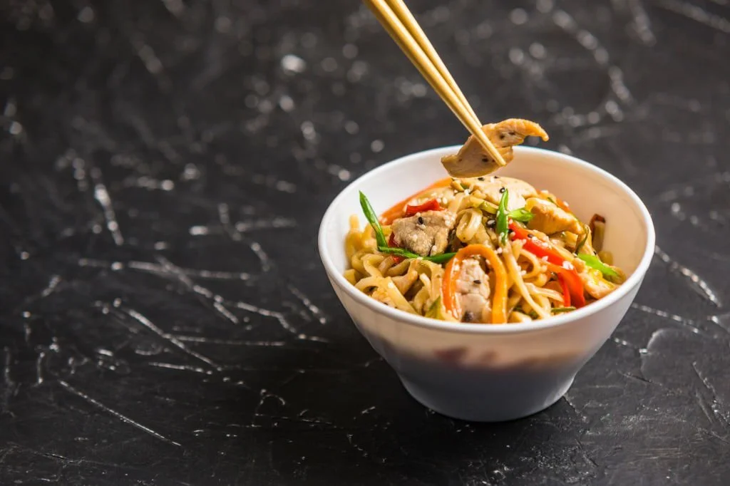 Chinese noodles in a Cup with chicken and vegetables with sticks. Wok food from the restaurant on a dark background from a side view.